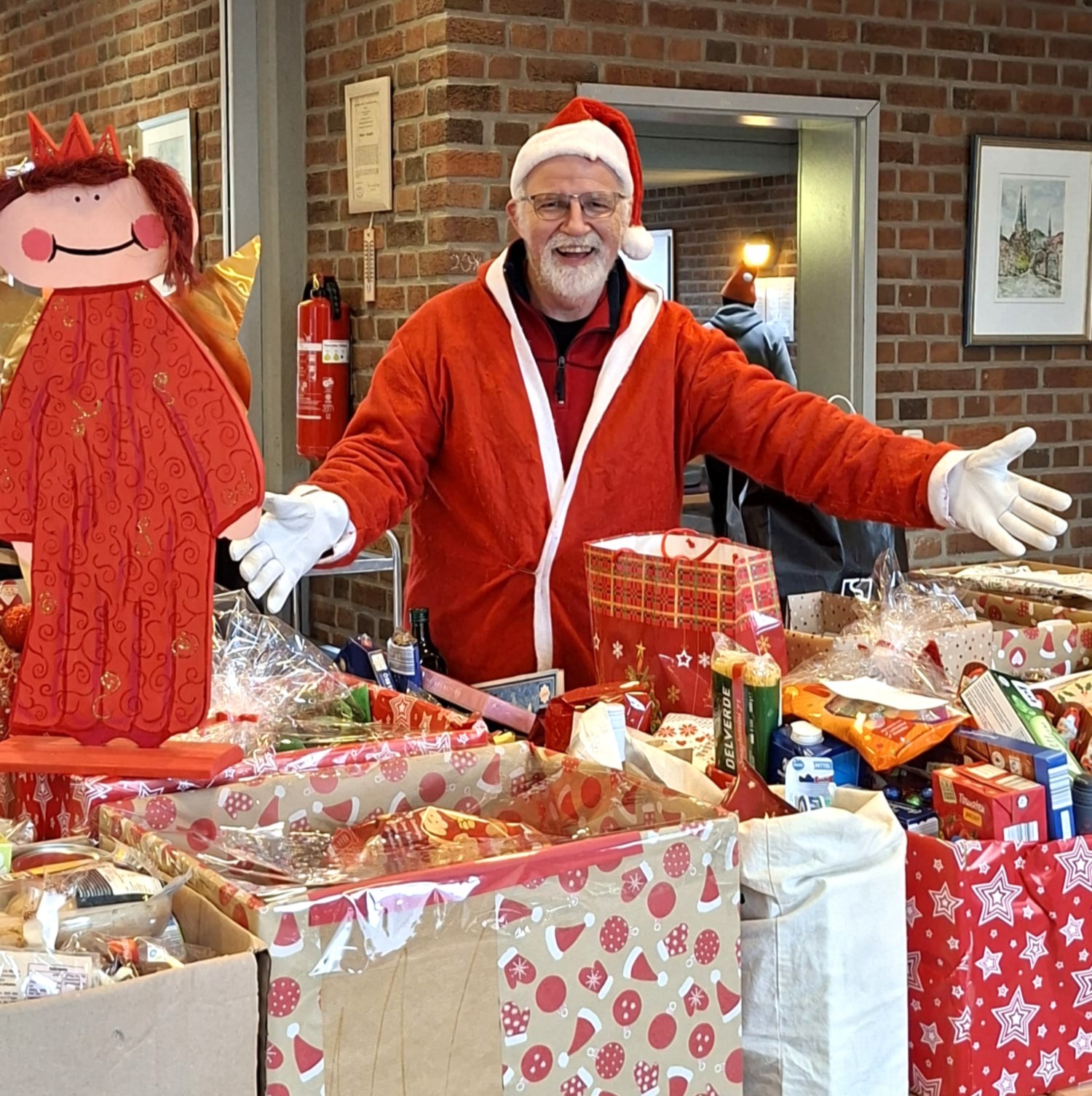 Ein älterer Mann im roten Weihnachtsmannkostüm lächelt fröhlich hinter Geschenken und Lebensmitteln