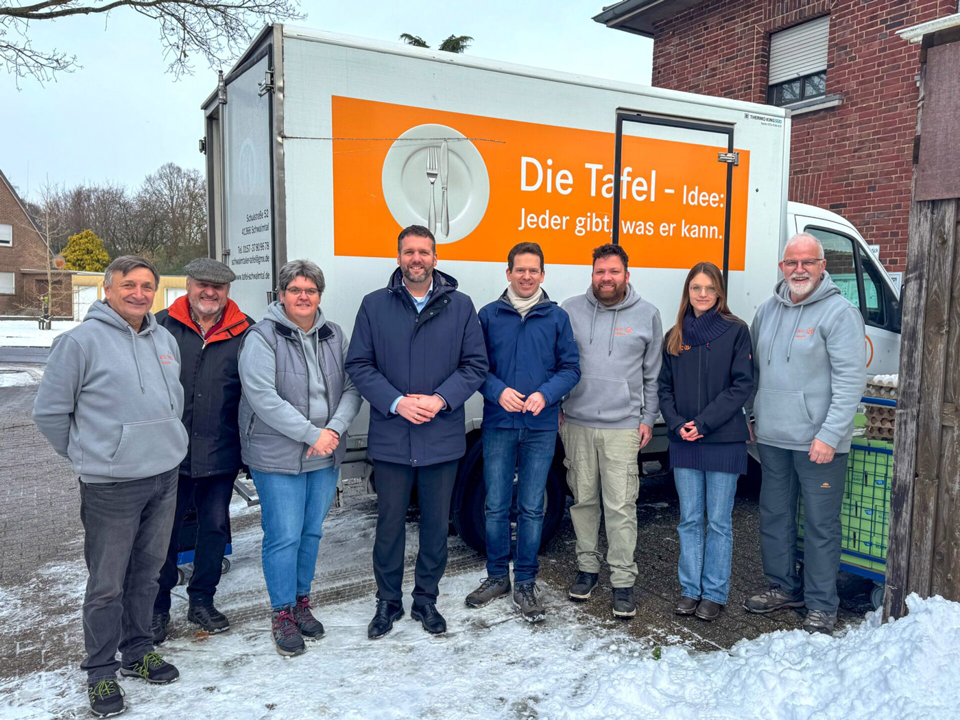 Acht Erwachsene stehen lächelnd im Schnee vor einem weißen Die Tafel Transporter