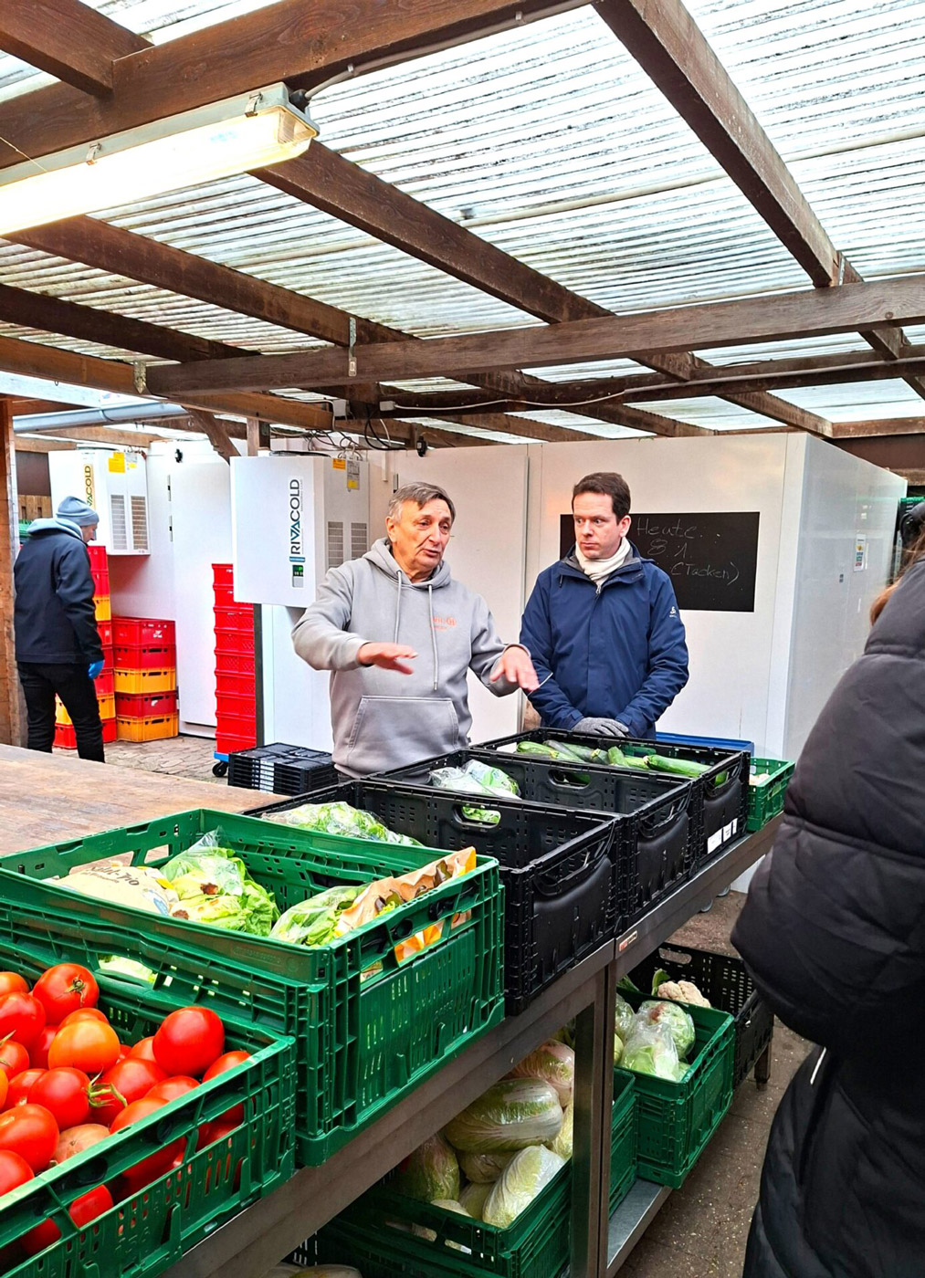 Zwei Männer unterhalten sich lebhaft an einem Gemüsestand mit frischem Gemüse in grünen Kisten